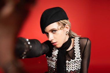 trendy nonbinary person in black beret looking at camera and posing with blurred outstretched hand on red background