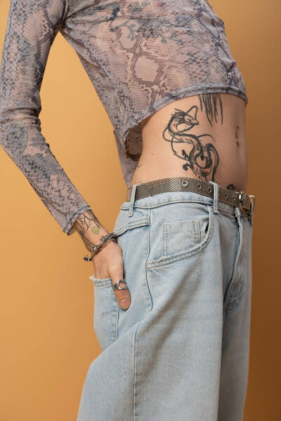 Cropped view of tattooed queer person holding hand in pocket of jeans on yellow background 