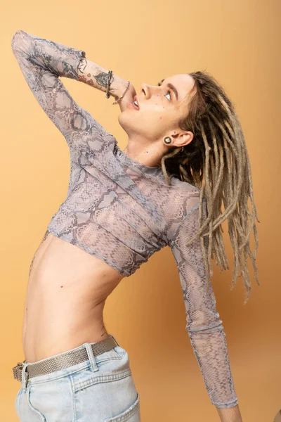 Young tattooed queer person with dreadlocks looking up on yellow background 