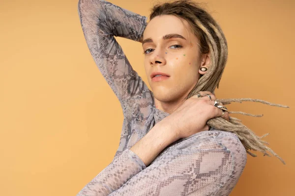 Portrait of tattooed queer person touching dreadlocks and looking at camera isolated on yellow 
