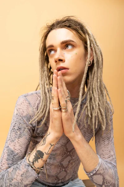 Tattooed queer person doing praying hands gesture on yellow background 