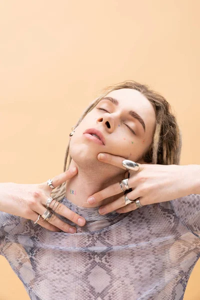Tattooed nonbinary person with silver accessories touching neck isolated on yellow 