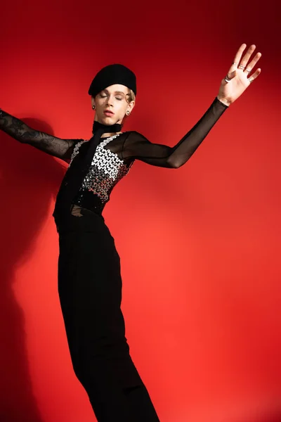 trendy nonbinary person in black skirt and beret standing with closed eyes and outstretched hands on red background with shadow
