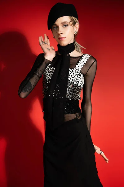 tattooed queer model in elegant attire waving hand and looking at camera on red background with shadow