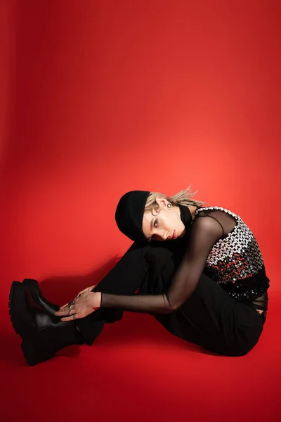 full length of nonbinary person in black beret and boots sitting at looking at camera on red background