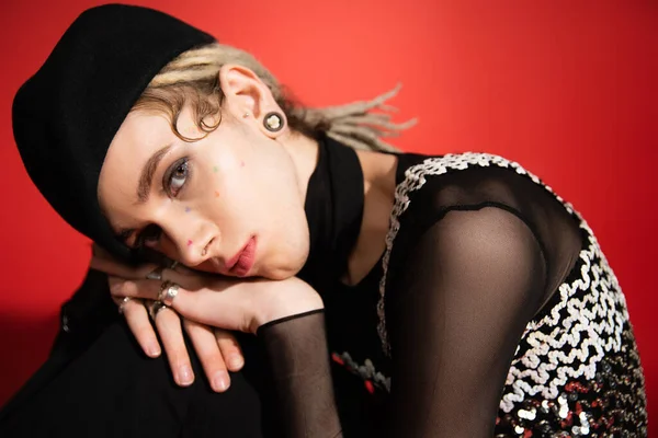 portrait of queer person in black beret and elegant top with sequins looking at camera on red background