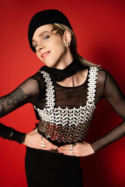 queer model in black beret and top with sequins holding hands on waist and looking at camera on red background