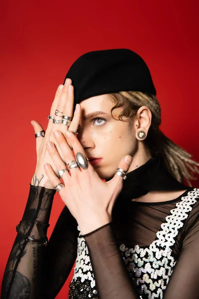 fashionable queer person in black beret and silver rings obscuring face with hands isolated on red