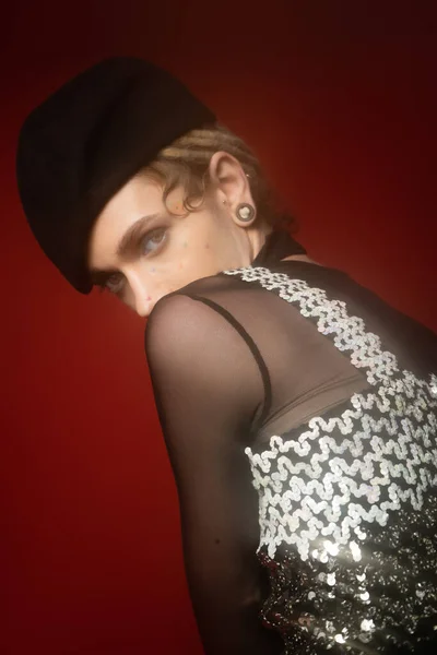 young nonbinary person in black beret and elegant top with sequins looking at camera on dark red background