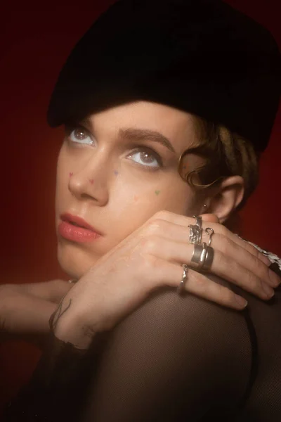 portrait of young queer model in silver rings and black beret looking away on dark red background