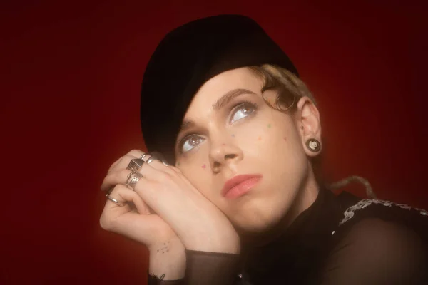 stock image portrait of young and stylish nonbinary person in black beret and silver rings looking away isolated on dark red