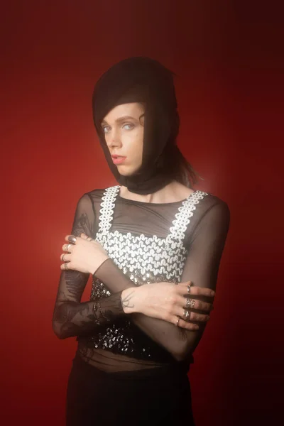 nonbinary person in black headwear and top with sequins posing with crossed arms on dark red background