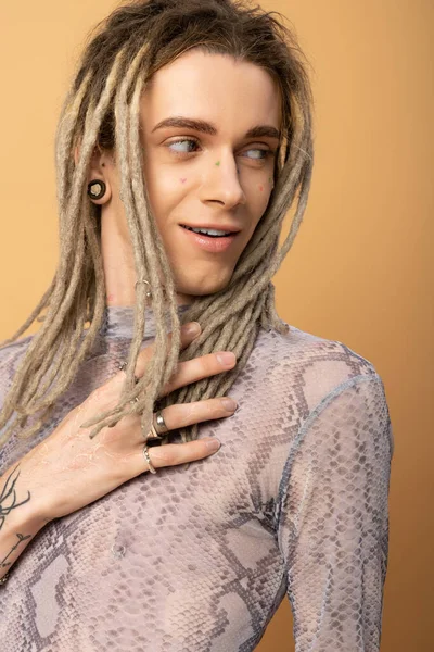 smiling queer person in top with snakeskin print touching dreadlock and looking away isolated on yellow