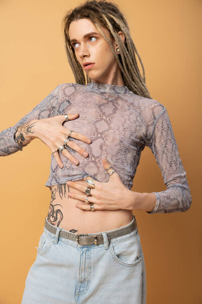 tattooed queer person with dreadlock posing in top with snakeskin print isolated on yellow