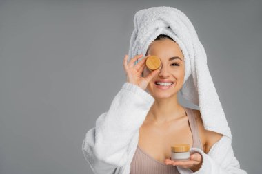 Smiling woman with towel on head holding cream near face isolated on grey 