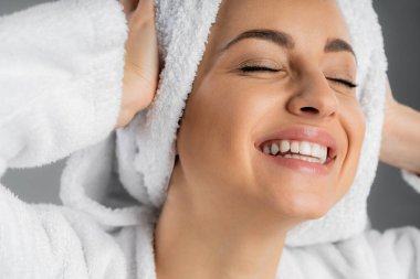 Cheerful young woman touching white towel on head isolated on grey 