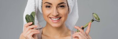 Cheerful woman with towel on head holding jade roller and gua sha isolated on grey, banner  clipart