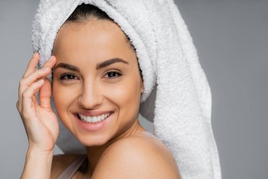 Portrait of smiling woman with towel on head touching face isolated on grey 