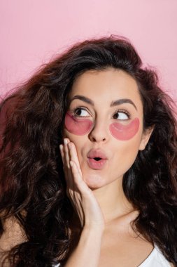 Dreamy woman with eye patches on face touching cheek on pink background 