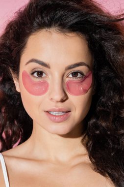 Portrait of curly young woman with hydrogel eye patches on pink background 