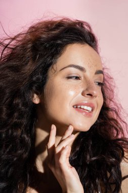 Positive young woman with freckles touching face isolated on pink  clipart