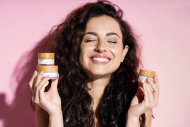 Positive freckled woman holding cosmetic creams on pink background 