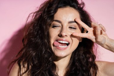 Young woman with freckles showing peace sign on pink background 