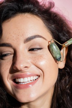 Close up view of smiling freckled woman using jade roller isolated on pink 