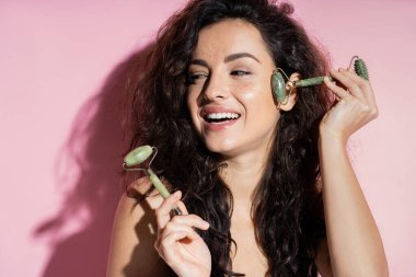 Cheerful freckled woman using jade roller on face on pink background  clipart