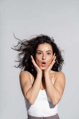 Excited curly woman in white top touching face isolated on grey 