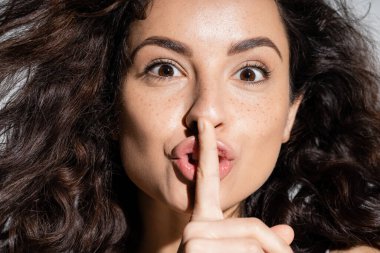 Portrait of curly freckled woman showing secret gesture isolated on grey 