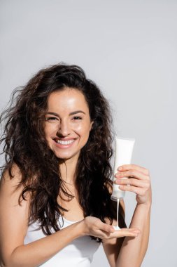 Smiling curly woman pouring body lotion on hand isolated on grey 