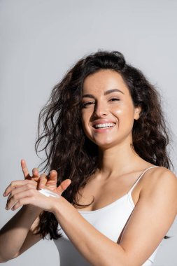 Cheerful woman in white top applying hand cream isolated on grey 