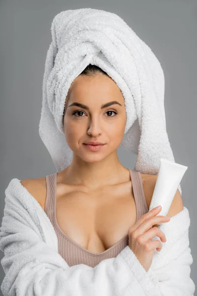 stock image Young woman with perfect skin and towel on head holding cream isolated on grey 