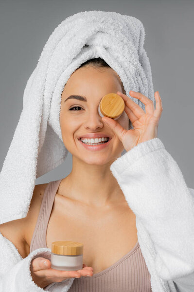 Positive woman with towel on head holding jars with cosmetic cream isolated on grey 