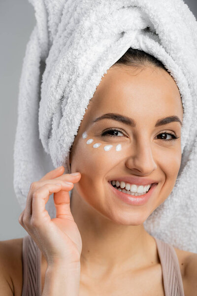 Positive woman with towel on head and cream on face looking at camera isolated on grey 
