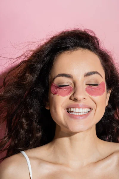 Smiling woman with hydrogel eye patches closing eyes on pink background