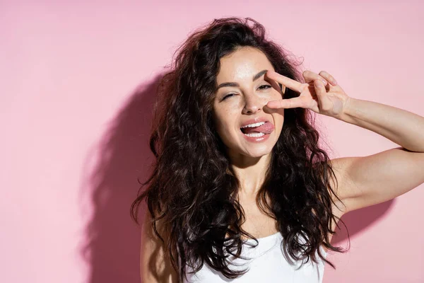 Young curly woman showing peace sign on pink background