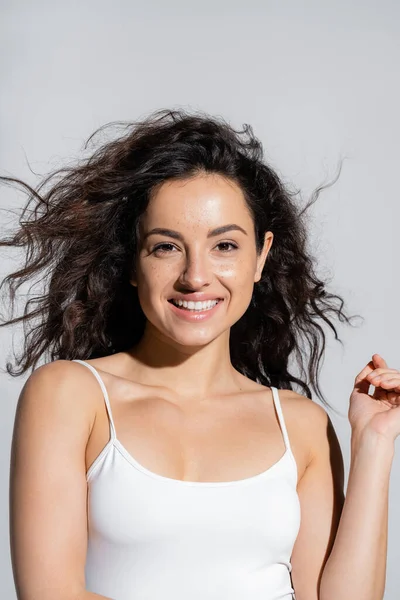 Portrait of cheerful curly woman in white top looking at camera isolated on grey 