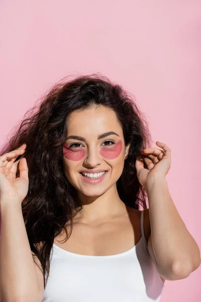 Cheerful young woman with hydrogel patches touching hair on pink background