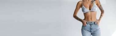 cropped view of slender woman in satin bra standing with hands in pockets of jeans on grey background, banner