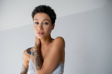 tattooed brunette woman in bralette touching neck and looking at camera on grey background
