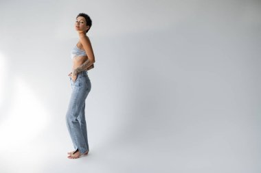 full length of barefoot brunette woman in bra and jeans standing with hand in pocket on grey background