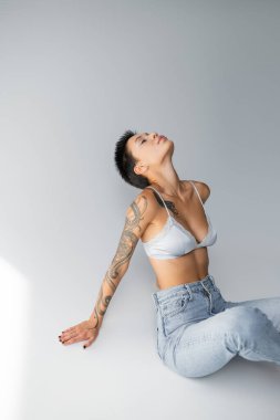 seductive tattooed woman in silk bra and jeans sitting with closed eyes on grey background