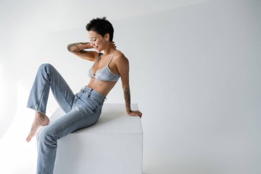 smiling brunette woman in silk bra and jeans sitting on cube on grey background