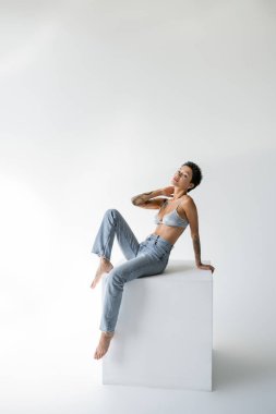 young barefoot woman in blue jeans and satin bra posing on cube and looking at camera on grey background