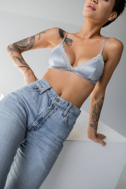 partial view of sexy woman in blue silk bralette and jeans posing with hand on hip on grey background