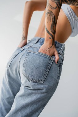 cropped view of tattooed woman in bra posing with hands in back pockets of blue jeans isolated on grey