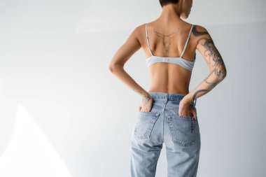 back view of tattooed woman in bra standing with hands in back pockets of jeans on grey background