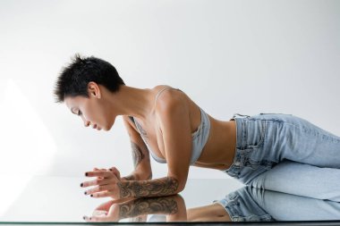 side view of brunette short haired woman in blue jeans and bra lying on mirror on grey background